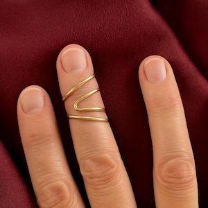 May include: A gold-colored ring with a unique design, featuring three connected bands that wrap around the finger. The ring has a minimalist style and is shown on a finger against a burgundy background.