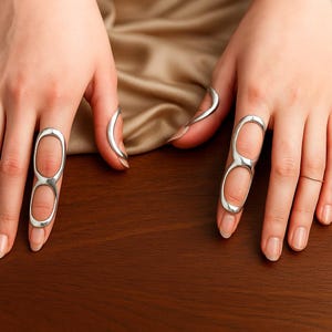 Puede incluir: Anillos de dedo plateados con un diseño alargado. Los anillos tienen una forma geométrica abierta, con dos aberturas ovaladas. Los anillos se llevan en los dedos, con un acabado pulido. El fondo es una superficie marrón oscuro.