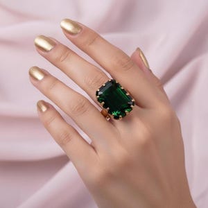 Puede incluir: Un anillo dorado con una gran piedra preciosa verde esmeralda de talla esmeralda. El anillo tiene una banda dorada y un engaste dorado decorativo alrededor de la piedra preciosa. El anillo se muestra en una mano.