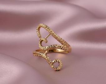May include: A gold-colored ring with an open design featuring tentacle-like shapes. The ring has a textured surface with small, round details. The ring is set against a soft, pink background.
