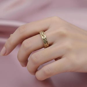 May include: A gold-colored ring with an intricate, swirling pattern. The ring is worn on a finger, showcasing its detailed design and polished finish. The ring's design features a series of interconnected, flowing shapes, creating a decorative band.