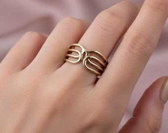 May include: A gold-colored ring with a unique open design. The ring features multiple parallel bands that curve and intersect, creating an eye-catching geometric pattern. The ring is worn on a finger, showcasing its elegant and modern style.