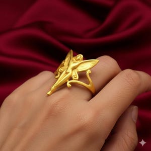 Può includere: Un anello dorato con un design a forma di fata con ali spiegate. L'anello è indossato su una mano, mettendo in risalto gli intricati dettagli della forma della fata. Il design dell'anello include curve delicate e una finitura lucida.