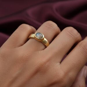 Puede incluir: Un anillo de oro con una piedra preciosa ovalada de color azul claro. El anillo tiene un diseño simple y elegante con una banda gruesa. La piedra preciosa está engastada en un engaste de bisel. El anillo se muestra en un dedo.