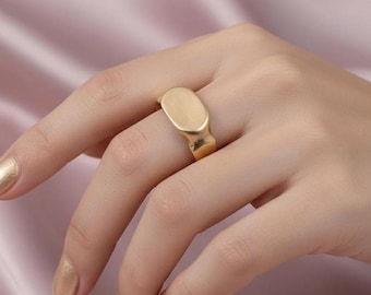 May include: A gold-colored signet ring with an oval face. The ring is worn on a finger against a soft pink background. The ring has a brushed metal finish and a classic, minimalist design.