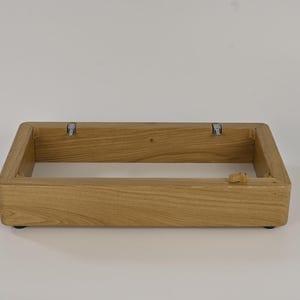 Könnte beinhalten: Ein rechteckiger Holzrahmen mit abgerundeten Ecken. Der Rahmen besteht aus hellfarbenem Holz und hat Metallhalterungen an der Innenseite. Kleine schwarze Füße sind unten sichtbar.