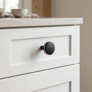 May include: A round cabinet knob featuring a matte black finish on the front face and a thin gold-toned metallic band around the side, mounted on a white drawer front. The hardware has a circular profile and a smooth, solid construction.
