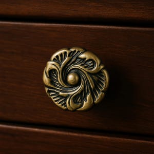 May include: Close-up of a bronze-colored cabinet knob with an ornate floral design. The knob is circular with a central sphere and swirling leaf-like patterns. It is mounted on a dark brown wooden surface.