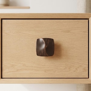 May include: A close-up of a wooden drawer with a dark brown, square drawer pull. The drawer is made of light-colored wood with a visible wood grain. The drawer pull has a curved design.