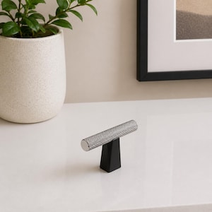 May include: A silver cabinet pull with a textured, cylindrical handle and a black, T-shaped base. The hardware is designed for furniture and has a modern aesthetic. The pull is approximately 4 inches long.
