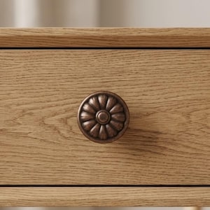 May include: Close-up of a wooden drawer with a decorative, bronze-colored knob. The knob has a floral design with a central circle and radiating petals. The wood grain is visible, showing a light brown color.
