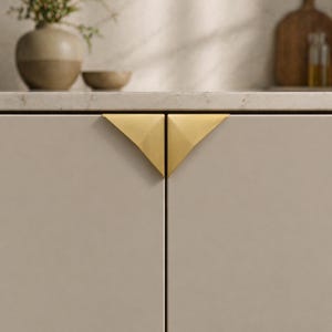 May include: Close-up of two cabinet doors with a light beige finish. Each door features a gold-colored, triangular handle. The handles are positioned to form a V-shape when the doors are closed. The cabinet is part of a kitchen setting.