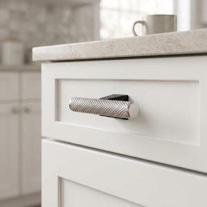 May include: A cylindrical cabinet pull with a silver-toned, cross-hatched knurled texture. The handle is mounted on a small, matte black rectangular base, which is attached to the center of a white, flat-panel wooden drawer front.