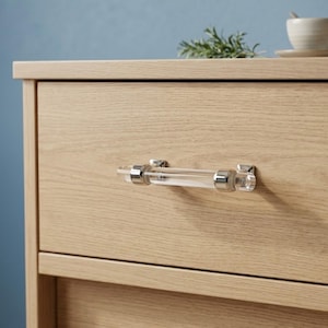 May include: A clear acrylic and chrome drawer pull is mounted on a light wood drawer. The cylindrical pull has chrome end caps and mounting hardware. The drawer is part of a light wood cabinet.