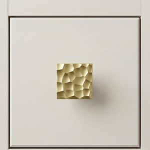 May include: A square, gold-toned cabinet knob with a textured, irregular surface. The knob is mounted on a white cabinet door, providing a contrast in color and texture. The hardware adds a touch of elegance to the furniture.