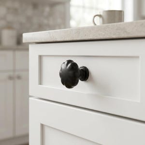 May include: A matte black cabinet knob with a rounded, scalloped, flower-like shape attached to a white drawer front. The knob features a smooth, curved surface with distinct ridges radiating from the center, providing a textured appearance.