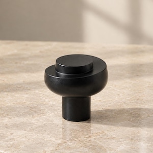 May include: A matte black cabinet knob featuring a cylindrical base and a wider, rounded top section with a smaller, raised circular disc centered on the upper surface. The hardware has a smooth, solid finish and a tiered, layered profile.