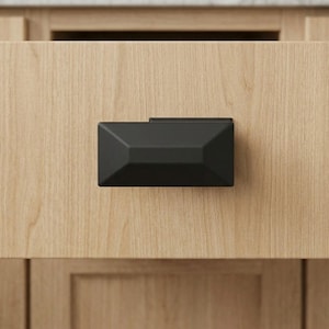 May include: A black, geometric cabinet pull is mounted on a light wood drawer front. The pull has a rectangular shape with angled sides and a matte finish. The wood grain is visible, adding texture to the design. The drawer is part of a larger cabinet.