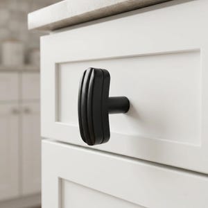 May include: A matte black cabinet knob with a vertical ribbed texture and a T-shaped profile. The hardware is mounted onto a white shaker-style drawer front, featuring a cylindrical base that connects the handle to the flat surface of the drawer.