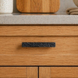 May include: Close-up of a black textured cabinet pull on a wooden cabinet. The pull is rectangular with a hammered metal appearance. The cabinet is made of light-colored wood with visible grain.