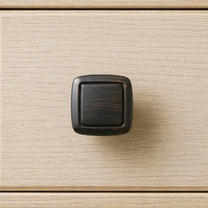 May include: A square, bronze-colored cabinet knob with rounded corners is centered on a light wood grain surface. The knob has a recessed square center and a slightly textured appearance, suggesting a metal construction.