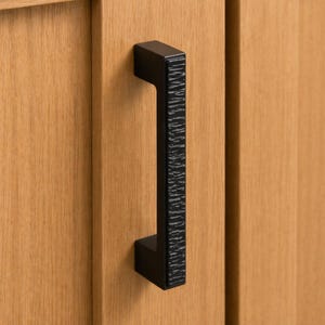 May include: Close-up of a black cabinet pull handle with a textured, vertical design. The handle is rectangular and mounted on a light-colored wooden cabinet door. The handle's textured surface adds a tactile element.