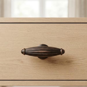 May include: A bronze-colored cabinet pull with a ribbed, oval design. The hardware is mounted on a light-colored wooden drawer, showcasing a classic and elegant aesthetic. The pull is centered on the drawer front.