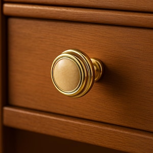 Puede incluir: Primer plano de un pomo de armario dorado con un centro beige claro. El pomo está fijado a un armario de madera con un acabado marrón cálido. El pomo tiene un diseño circular con anillos concéntricos.