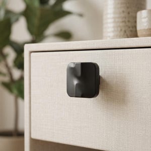 May include: A black, square drawer pull with a unique, sculpted design. The hardware is mounted on a light beige drawer front with a textured fabric appearance. The background includes a potted plant and decorative pottery.