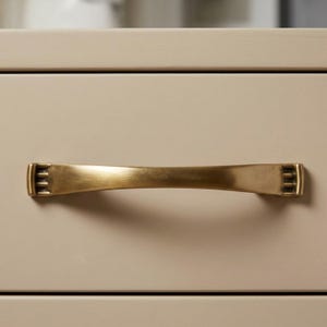 May include: A close-up of a brass-colored drawer pull with a curved handle and rectangular end details. The pull is mounted on a light beige cabinet drawer, showcasing a simple, yet elegant design. This hardware is suitable for various furniture styles.