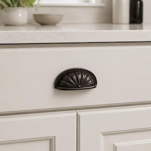 May include: A dark bronze, half-moon shaped cabinet pull with a detailed, fan-like design. The pull is mounted on a white cabinet door. The hardware adds a decorative touch to kitchen or bathroom cabinetry.