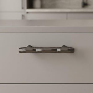 May include: A close-up of a gray cabinet drawer with a geometric, dark gray handle. The handle has a faceted design and is mounted horizontally. The drawer and handle are set against a neutral background, suggesting a modern kitchen or bathroom.