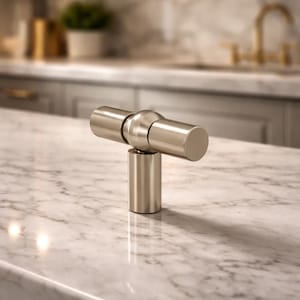 May include: A brushed nickel cabinet knob with a cylindrical handle and a round base. The hardware is set against a marble countertop, suggesting a kitchen or bathroom setting. The knob's design is modern and minimalist.