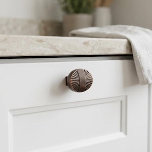 May include: A round, copper-toned metal cabinet knob featuring a textured design with vertical lines radiating from a central band. The knob is mounted on a smooth, white cabinet door with a recessed panel detail, providing a clear view of the hardware.