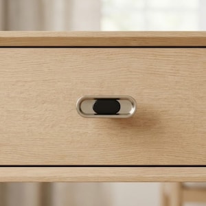 May include: Close-up of a light-colored wooden drawer with a silver and black oval-shaped handle. The handle is centered on the drawer front, providing a modern and minimalist aesthetic. The wood grain is visible.