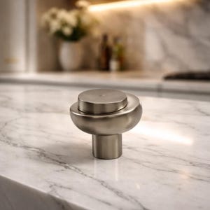 May include: A brushed nickel cabinet knob with a mushroom-shaped design. The knob features a circular top and a rounded base, mounted on a cylindrical stem. The hardware is set against a marble countertop in a kitchen setting.