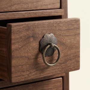 May include: Close-up of a wooden drawer with a decorative bronze pull. The drawer is open, revealing the interior. The wood has a rich, dark brown color and visible grain. The pull is a ring attached to a floral-shaped plate.