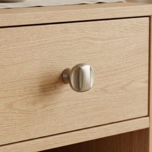 May include: Close-up of a light-colored wooden drawer with a brushed metal knob. The knob is oval-shaped and mounted on the drawer front. The wood grain is visible, and the drawer is part of a larger piece of furniture.