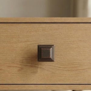 May include: Close-up of a square, bronze-colored cabinet knob. The knob is mounted on a light-colored wooden drawer, showcasing the wood grain. The knob has a raised, beveled design.