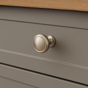 May include: A close-up of a brushed nickel cabinet knob with a round, ridged face. The knob is mounted on a gray cabinet door with a wooden top. The hardware has a classic, timeless design.