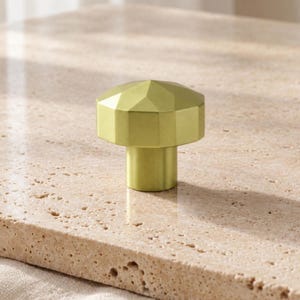 May include: A matte gold cabinet knob featuring a faceted, geometric top with multiple angled surfaces and a cylindrical base. The hardware has a smooth, metallic finish and a distinct multi-sided shape that creates a structured appearance.