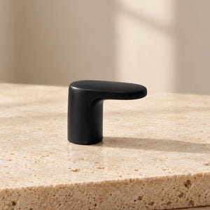 May include: A matte black cabinet knob featuring a cylindrical base and a horizontal, slightly rounded protruding handle. The hardware has a smooth, solid finish and a distinct L-shaped profile, designed for attachment to furniture or doors.