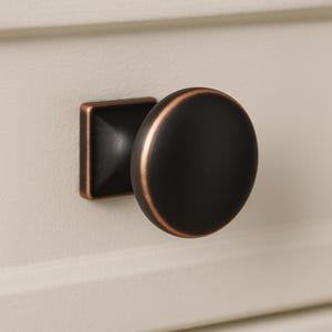 May include: Close-up of a cabinet knob with a dark, matte black finish and a copper-colored rim. The knob has a rounded, circular shape and is attached to a square base with a matching copper finish. The hardware is mounted on a cream-colored surface.