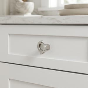 May include: A brushed silver cabinet knob with a circular, split-ring design mounted on a white wooden drawer front. The hardware features a smooth, metallic finish and a geometric, open-center shape that is centered on the drawer panel.