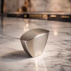 May include: A brushed nickel cabinet knob with a geometric design. The knob has a rounded top and a trapezoidal shape, with a smooth, metallic finish. It is set against a marble countertop.