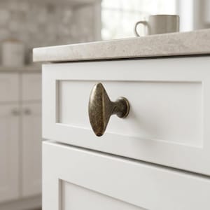 May include: An oval-shaped cabinet knob with a dark, weathered bronze finish attached to a white drawer front. The knob features a smooth, elongated design with a central stem that connects to the flat surface of the white cabinetry drawer.