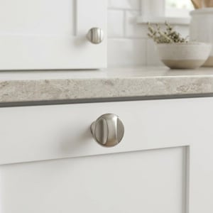 May include: A brushed nickel cabinet knob with a rounded, pill-shaped design featuring a central vertical indentation. The hardware is mounted on a white cabinet drawer front, showcasing a smooth metallic finish and a modern, clean aesthetic.