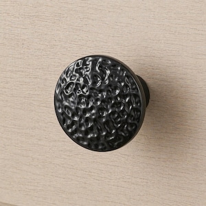 May include: A black, textured cabinet knob with a round face and a dark finish. The knob is mounted on a light-colored wooden surface, providing a contrast in color and texture. The knob's surface has a hammered appearance.