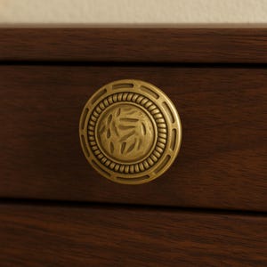 May include: A round, antique brass drawer pull with a detailed leaf design in the center. The hardware is set against a dark brown wooden surface, creating a contrast in color and texture. The pull has a decorative, vintage aesthetic.