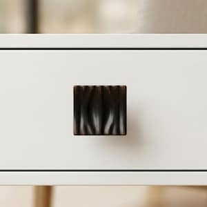 May include: A square, dark brown cabinet knob with a textured, wavy surface. The knob is centered on a white cabinet door, providing a striking contrast. The hardware adds a touch of rustic elegance to the furniture.